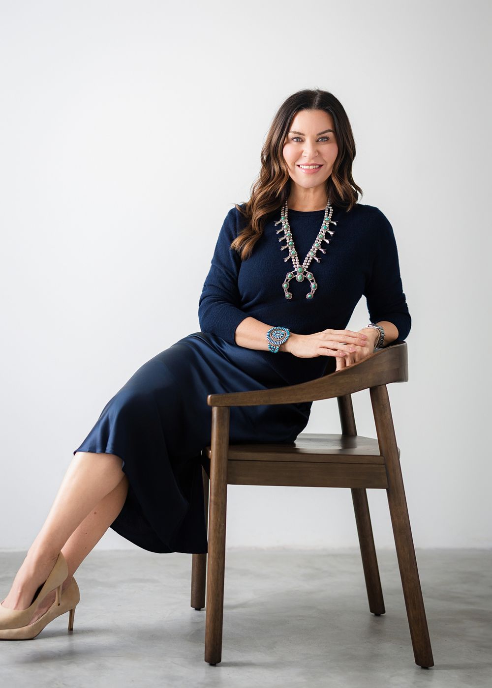 Woman in navy dress and squash blossom necklace seated on wooden chair against white background.