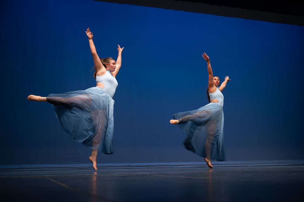 Two female ballet dancers in flowing blue dresses perform an arabesque pose together on a blue-lit stage.