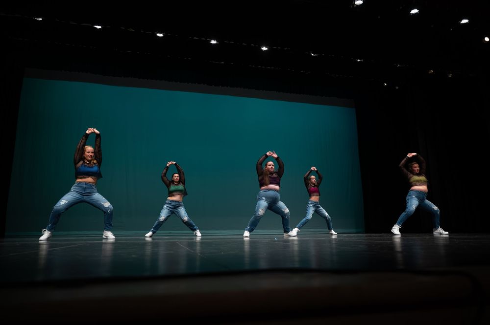 Five dancers in jeans and colorful tops perform a synchronized hip-hop routine on a teal-lit stage.
