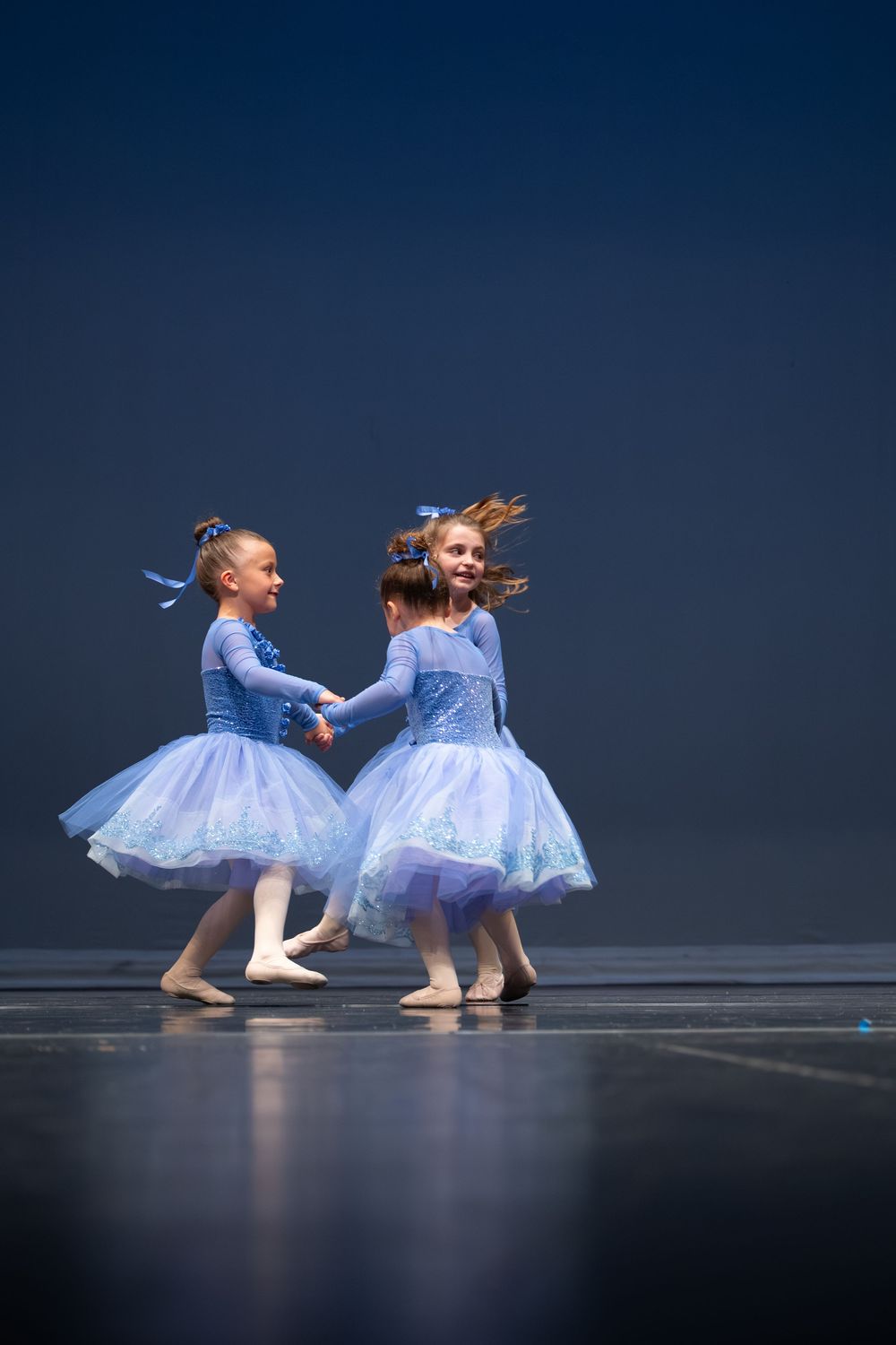 Three young girls in blue ballet tutus hold hands and spin together on a dark stage during a dance recital.