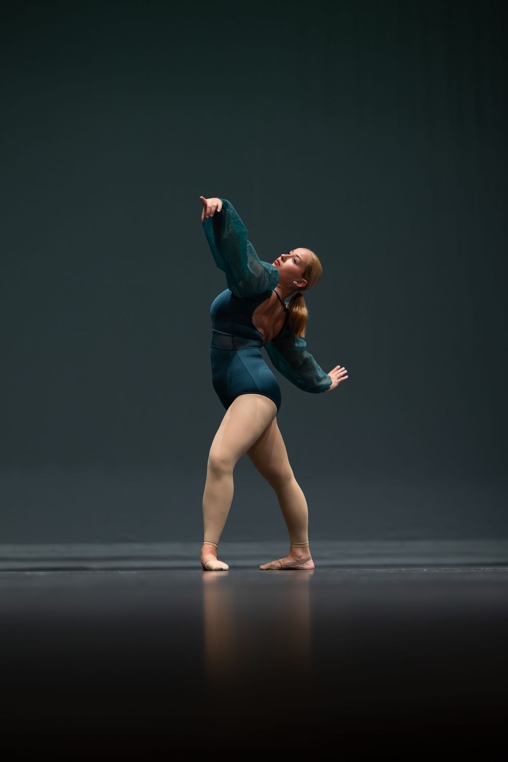 Female dancer in teal leotard performs a dramatic contemporary dance solo on a dark stage.