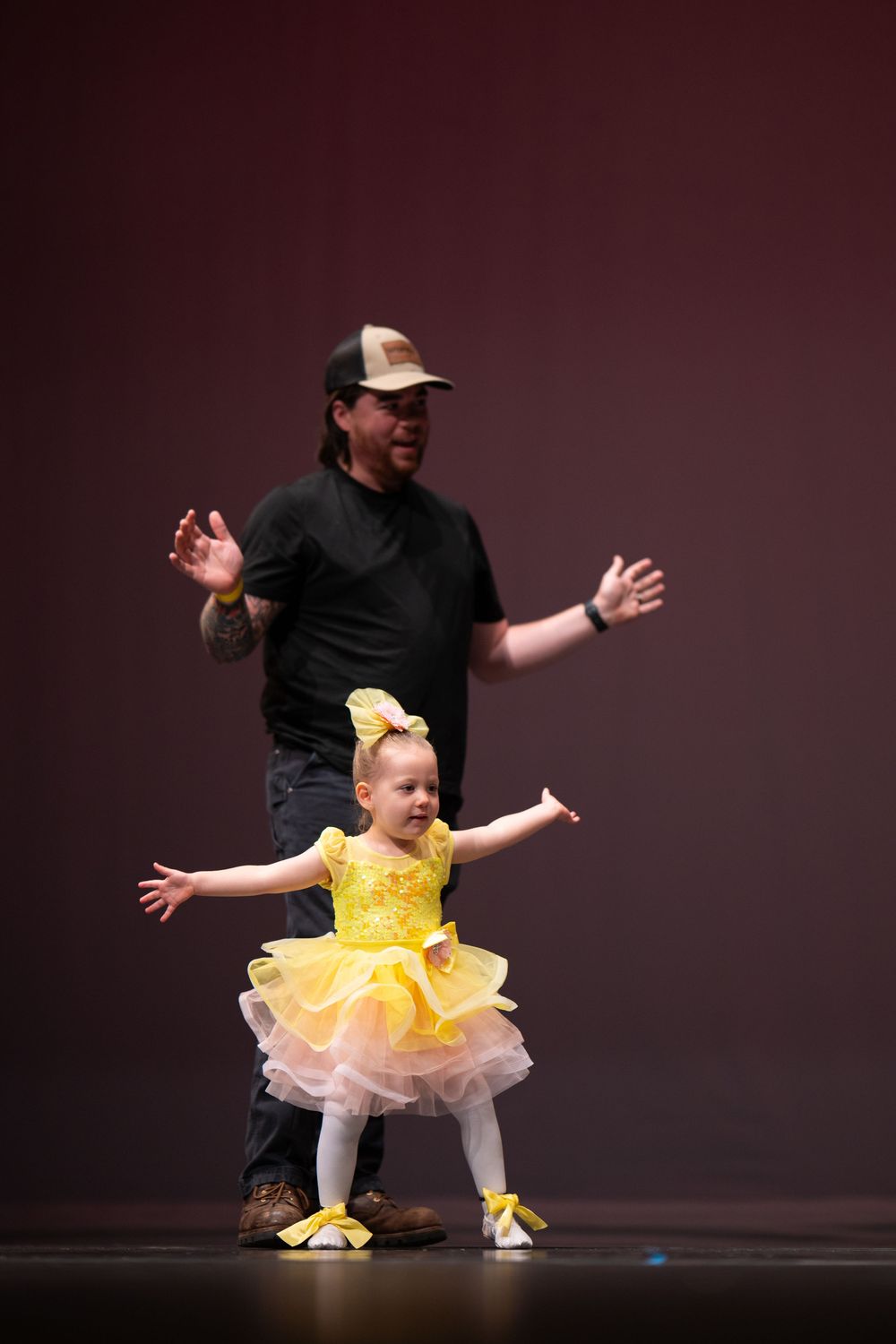 A smiling toddler girl in a yellow tutu dress performs on stage with her father behind her at a dance recital.