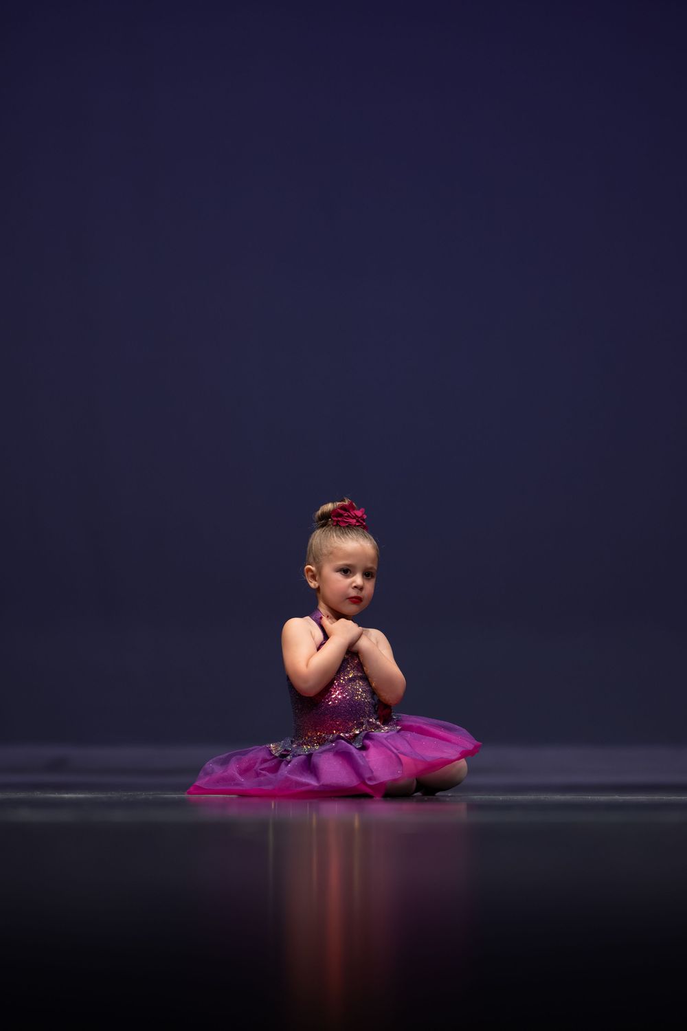 Adorable toddler in pink tutu sits on stage floor with hands clasped during ballet recital.