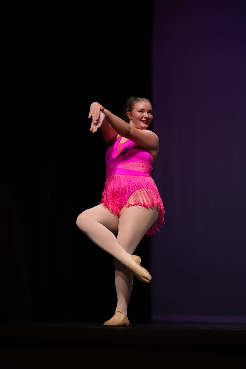 Teen dancer in bright pink fringed costume strikes a pose on a dimly lit performance stage.