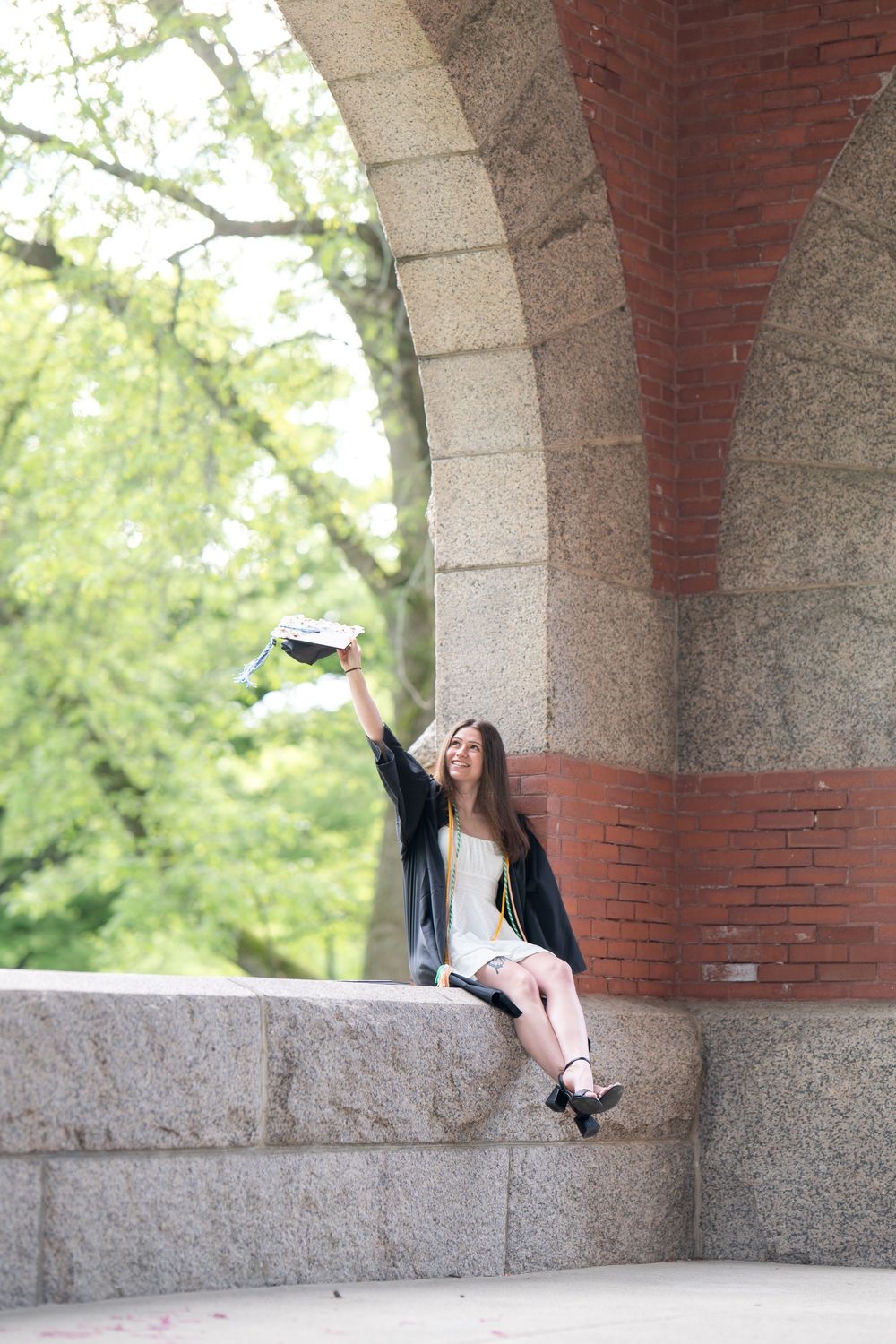 Graduate in black robe and white dress raises her cap triumphantly while sitting under a stone archway.