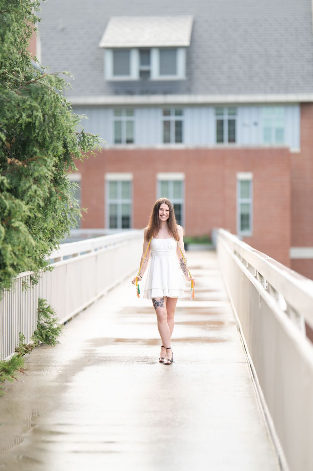 Smiling young woman in white dress walks across a bridge holding graduation tassels near a brick building.