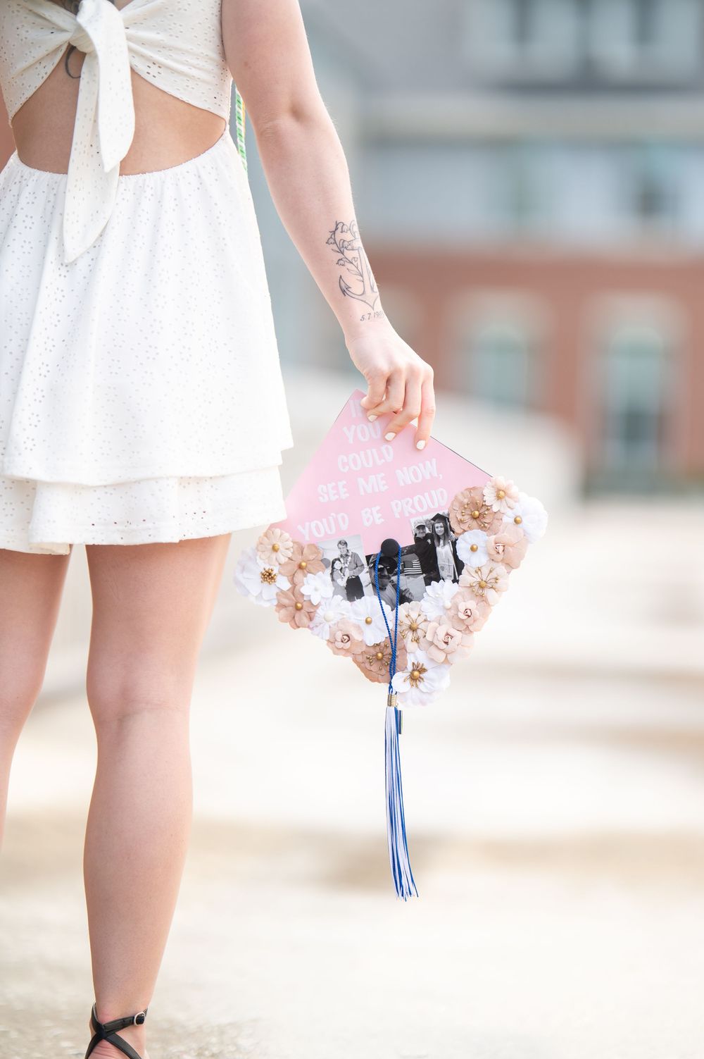 Graduate in white dress holds a decorated floral pink graduation cap with photos and blue tassel.