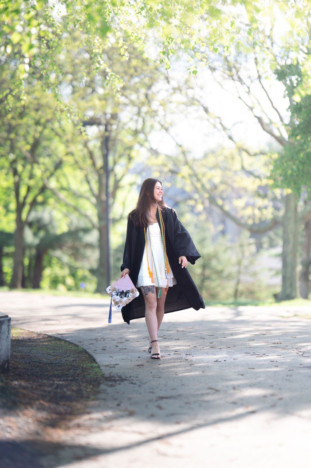 Smiling graduate walks in black cap and gown through a sunny tree-lined campus pathway.