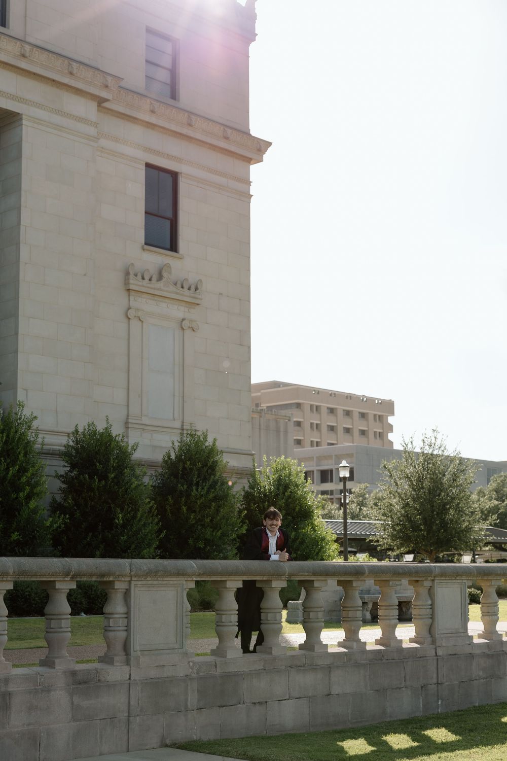Person standing near ornate stone balustrade outside a grand neoclassical government building with manicured shrubs.
