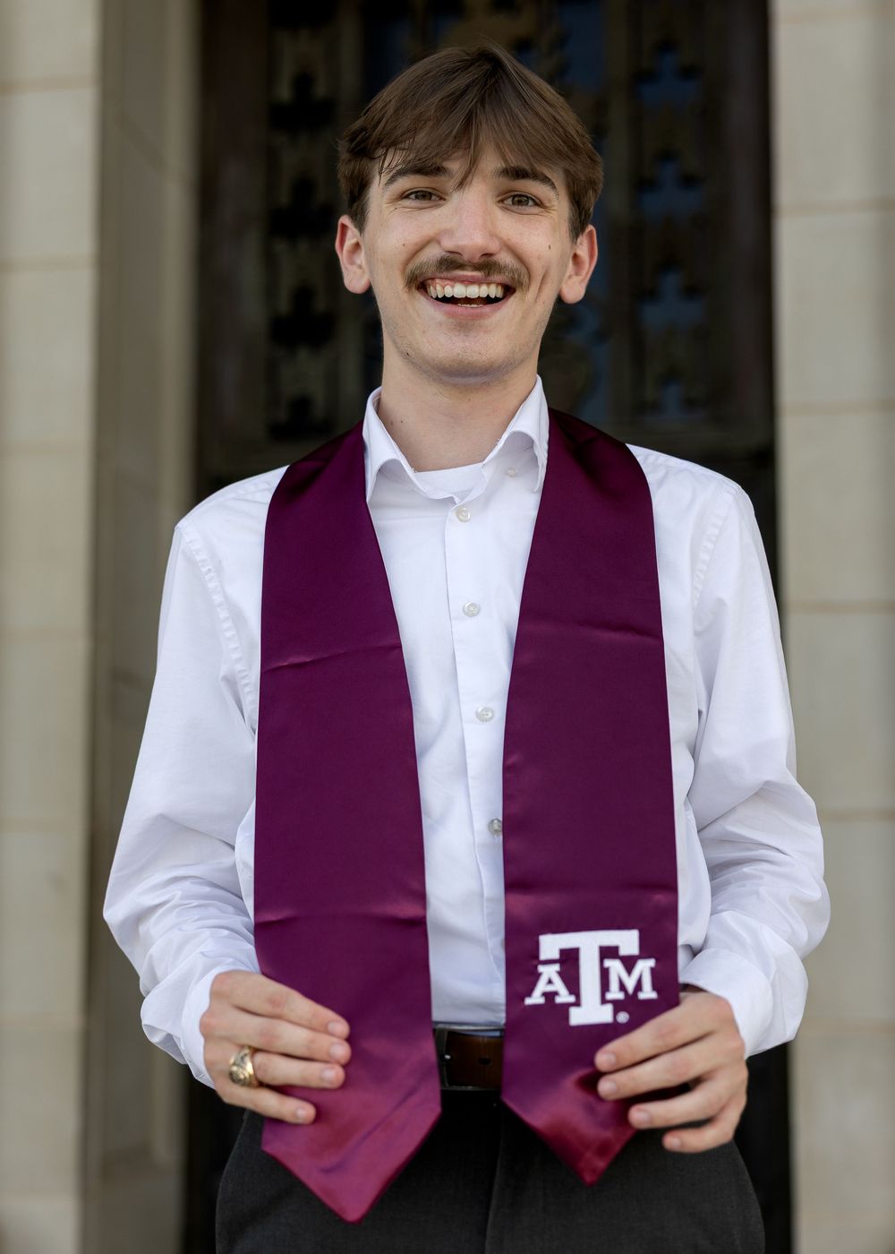 Smiling young man holding a maroon Texas A&M University graduation stole, wearing a white dress shirt.