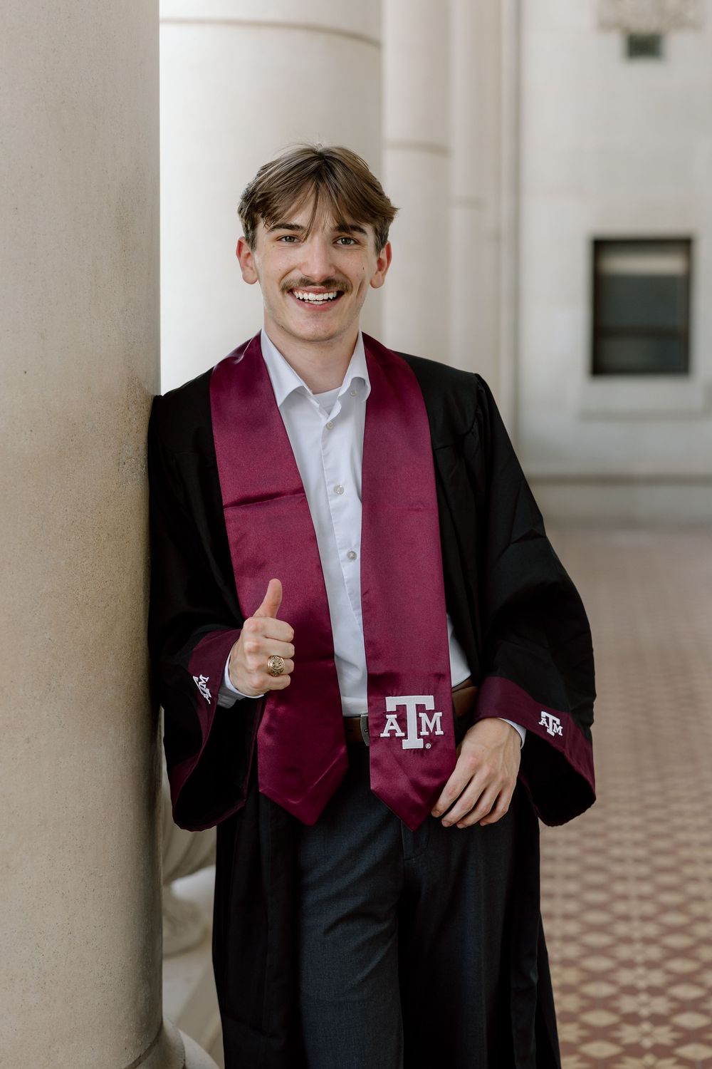 Texas A&M graduate in black gown and maroon ATM stole giving thumbs up while leaning against column.