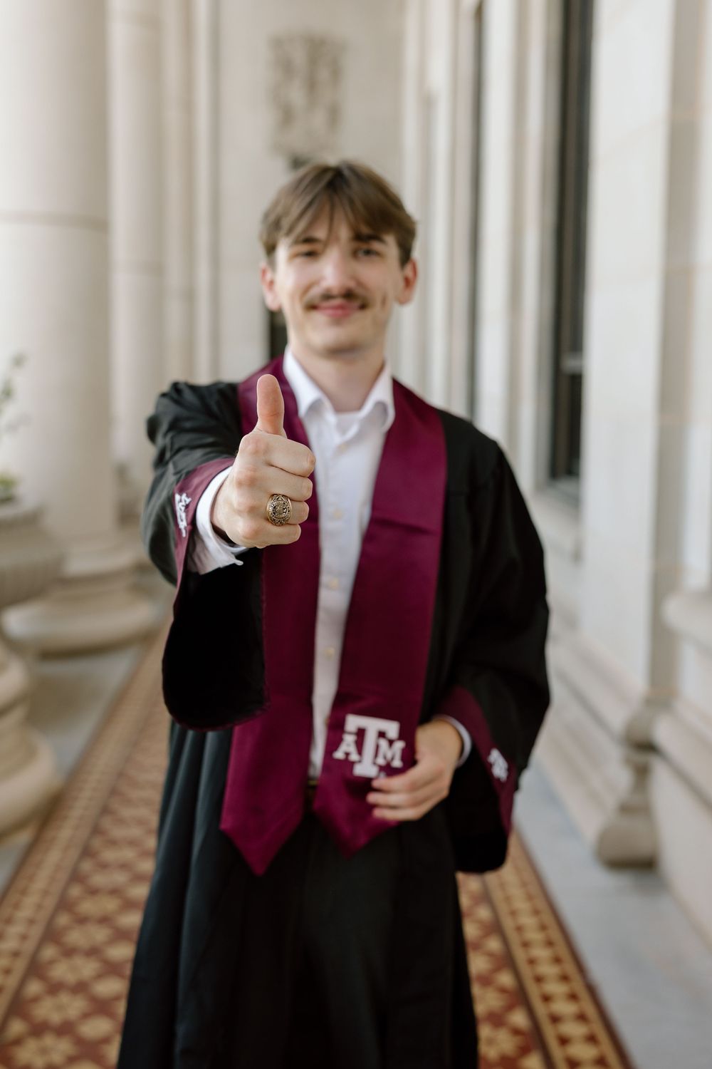 Texas A&M graduate in black gown and maroon stole gives thumbs up while wearing Aggie ring.