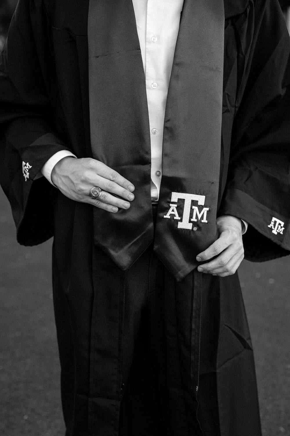 Black and white photo of Texas A&M graduate in cap and gown holding ATM stole, wearing class ring.