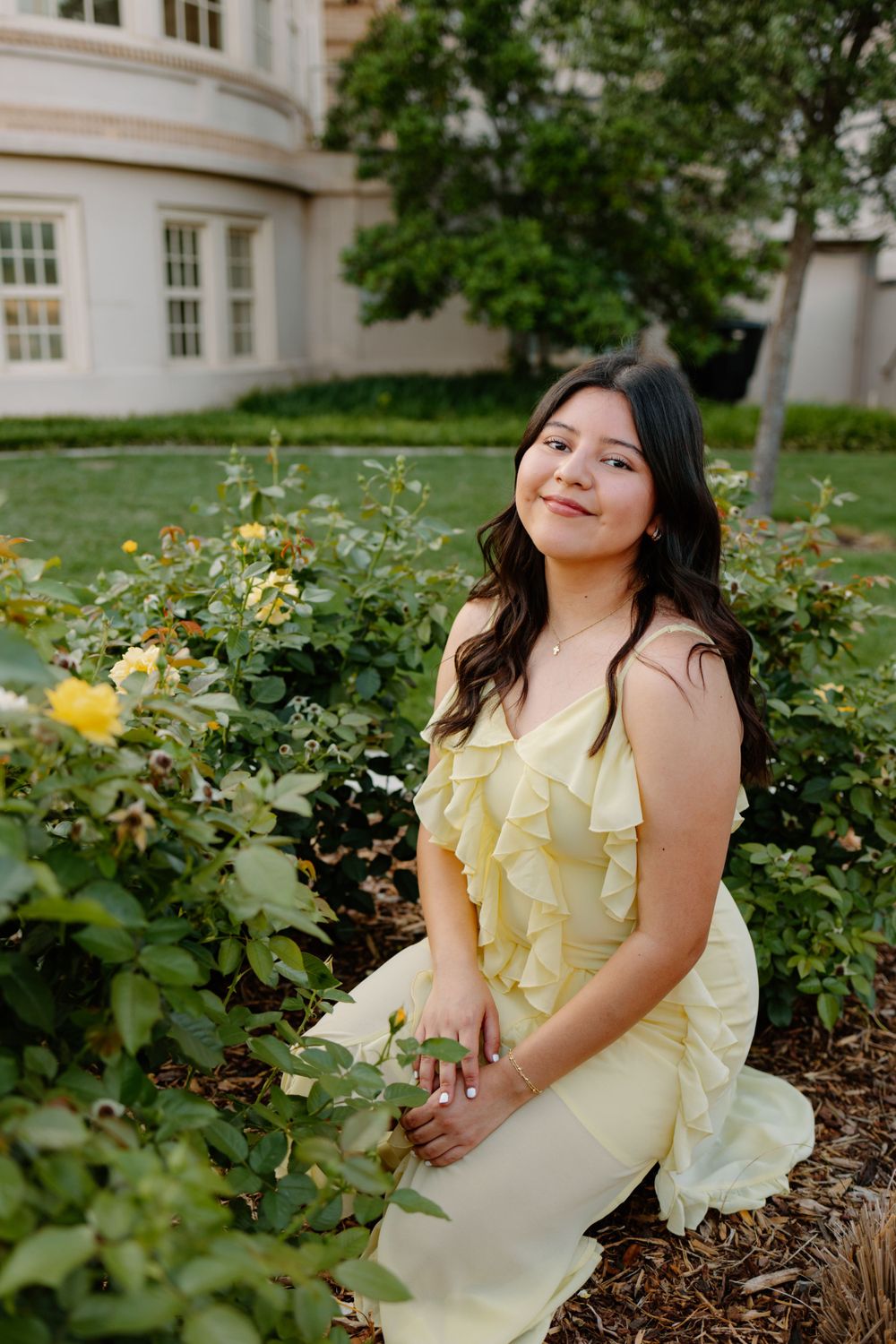Person in pale yellow dress poses by blooming yellow roses in garden setting.