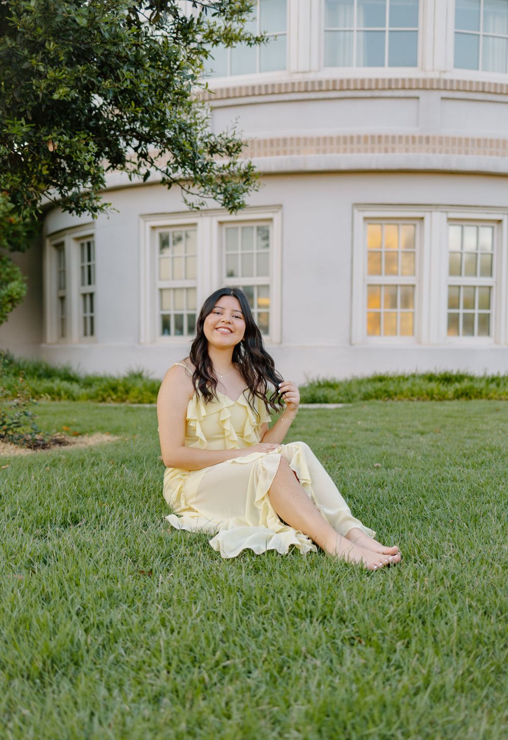 A person in a pale yellow dress sits smiling on a grassy lawn in front of a white building.