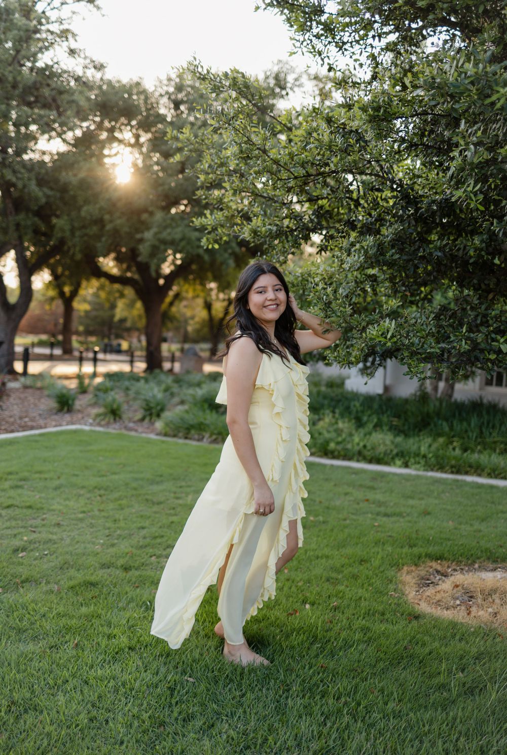 A person poses in a flowing yellow outfit against a sunset garden backdrop.