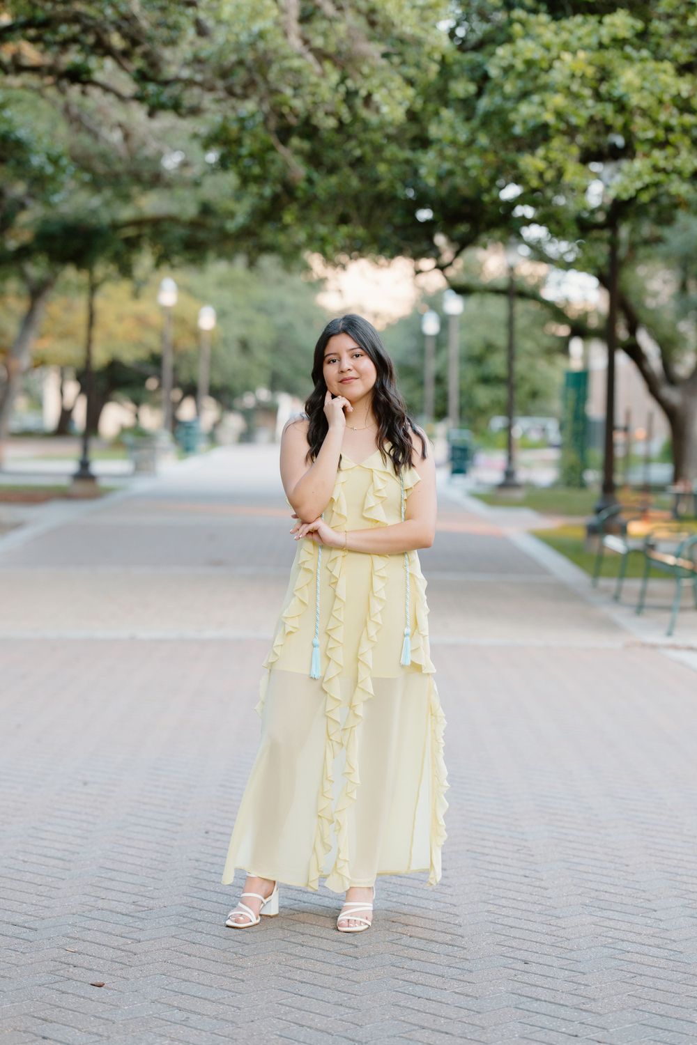 Person in yellow flowy dress poses on tree-lined campus pathway at golden hour.