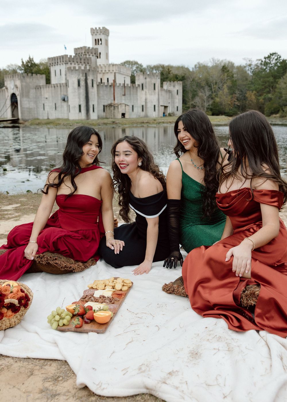 Four women in elegant gowns enjoy a picnic by a castle lake, smiling together on a white blanket.