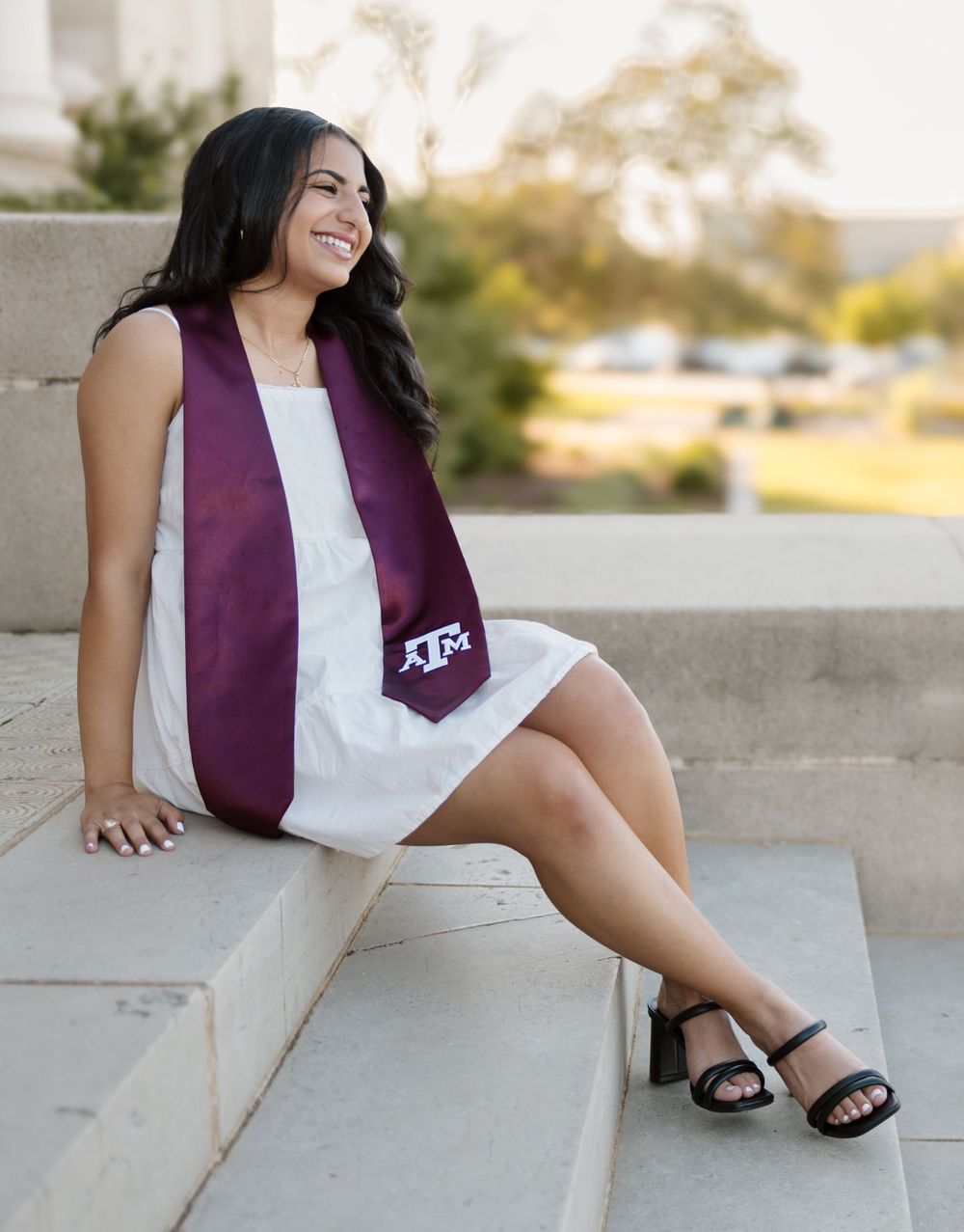 Graduate in white dress with maroon stole sits on concrete steps during graduation photoshoot.