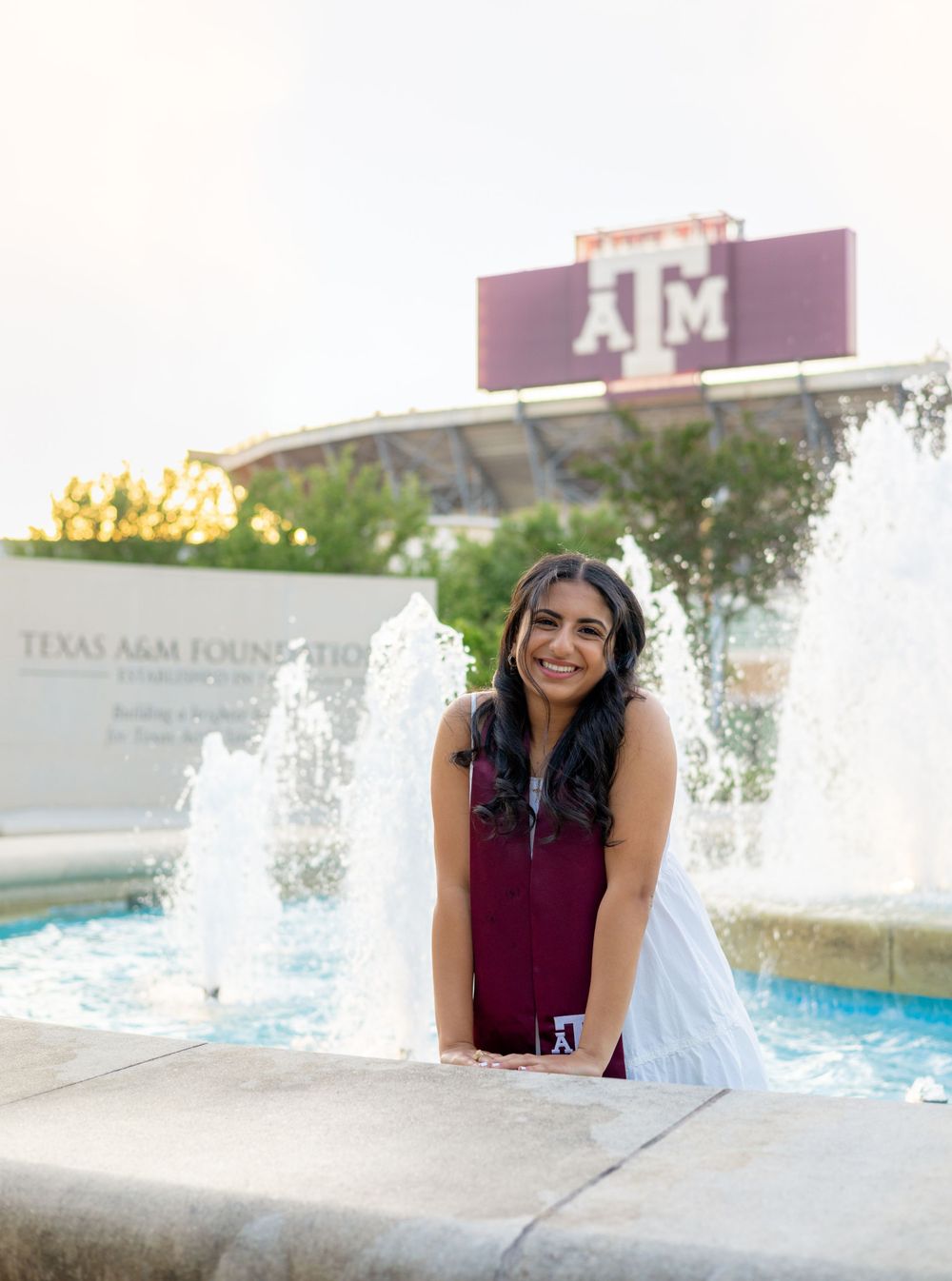 Graduate poses by fountain with Texas A&M sign visible in background during golden hour.