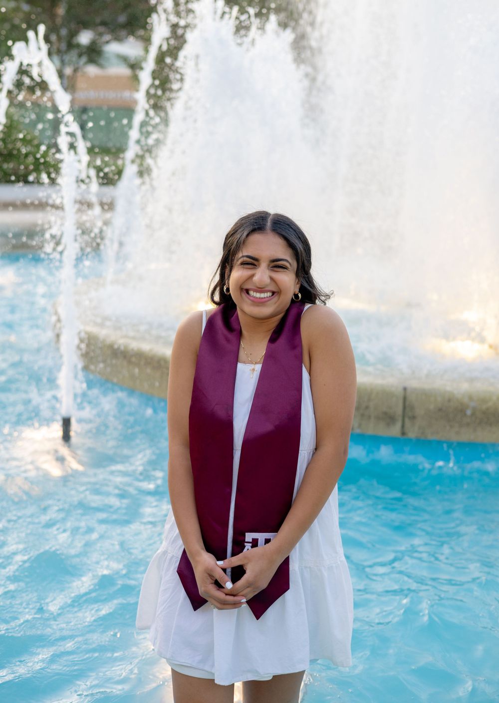 Graduate in white dress and purple stole smiles by fountain at Texas A&M campus during sunset celebration.