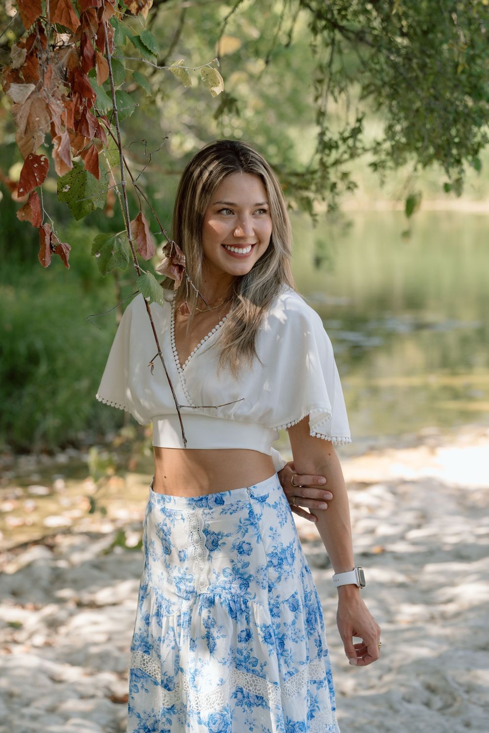 Smiling woman in white crop top and blue floral skirt standing near a river surrounded by trees.