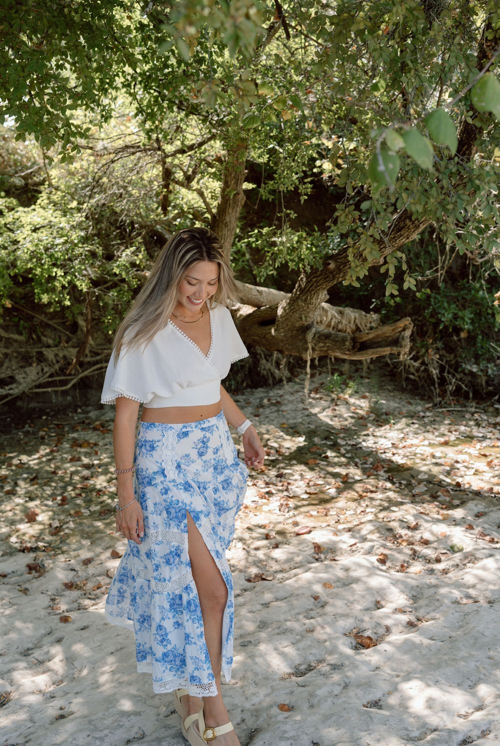 Woman in white crop top and blue floral maxi skirt smiling outdoors near trees on sandy ground.