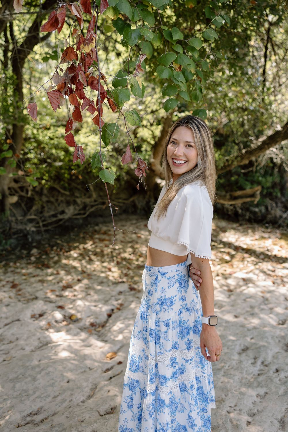 Smiling woman in white crop top and blue floral maxi skirt posing outdoors near lush green trees on sandy ground.