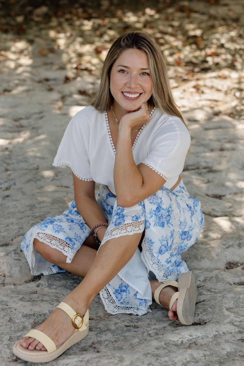 Smiling woman in white top and blue floral skirt sitting on sandy ground wearing sandals.