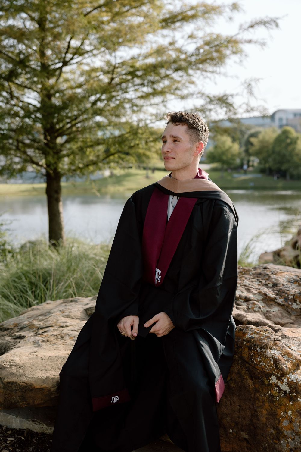 Young graduate in black and maroon academic gown sits on rocks near a lake, gazing thoughtfully into the distance.