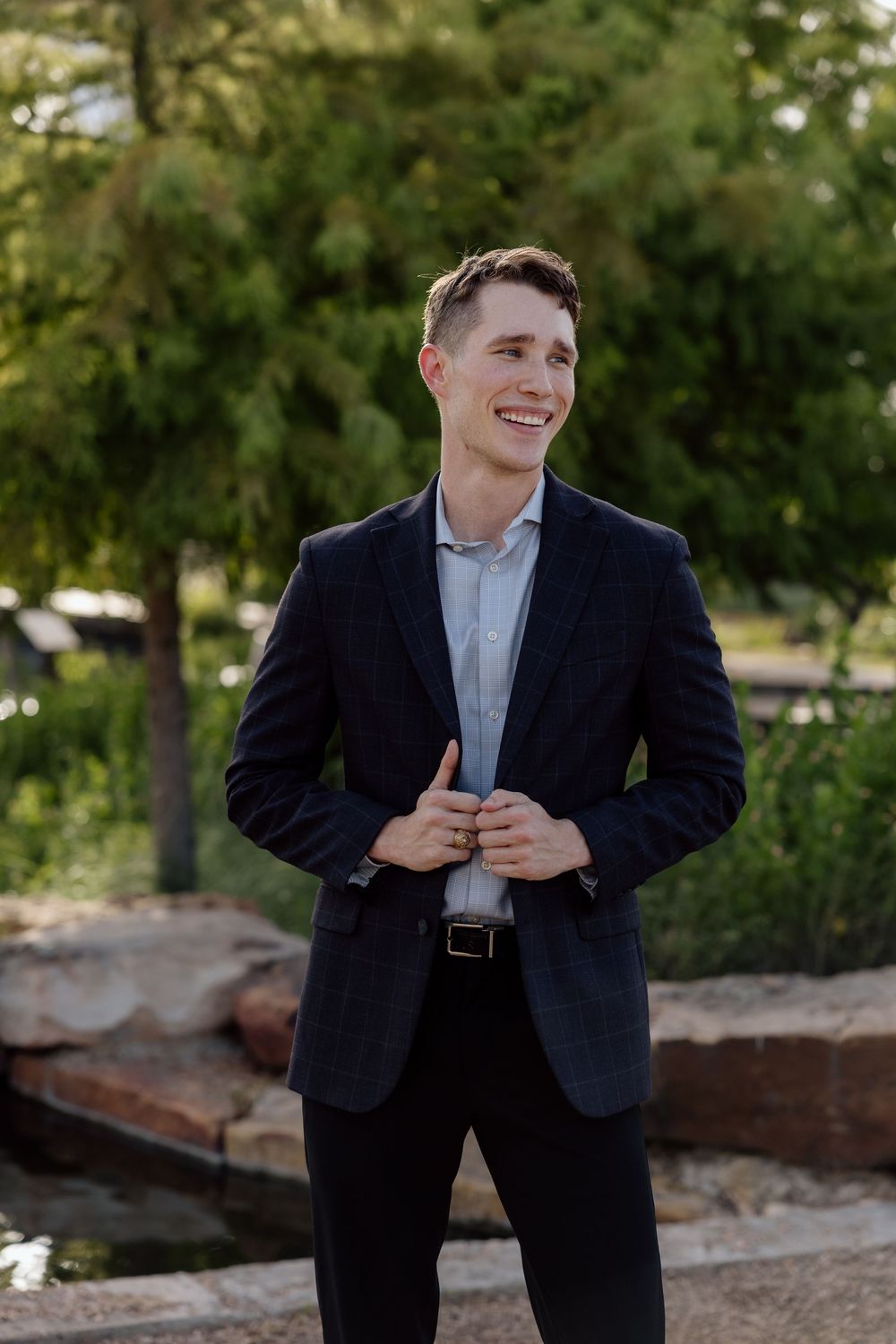 Smiling young man in navy suit adjusting jacket outdoors with green trees in background.