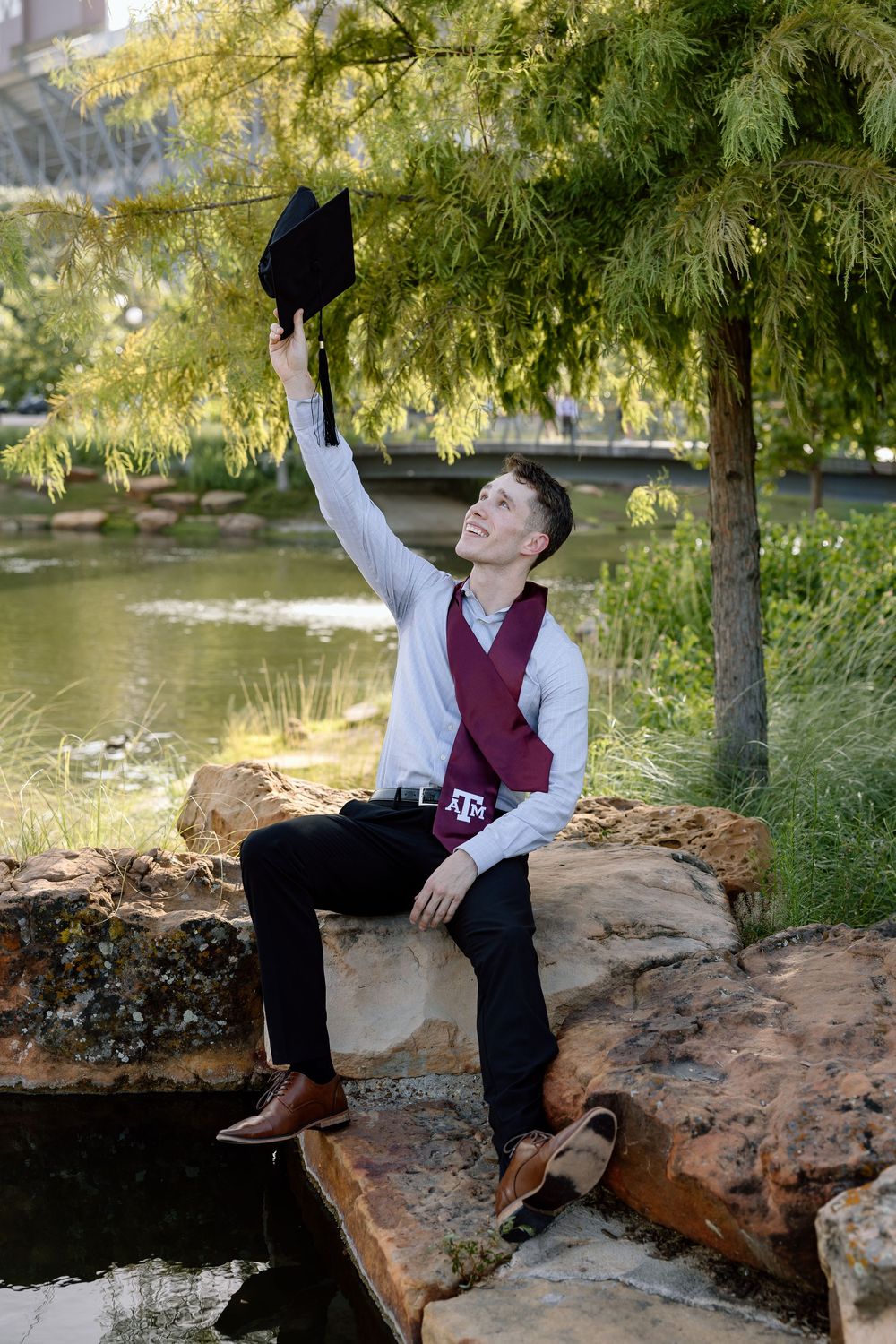A young man in maroon vest and sash celebrates graduation by raising his cap near a pond at Texas A&M University.