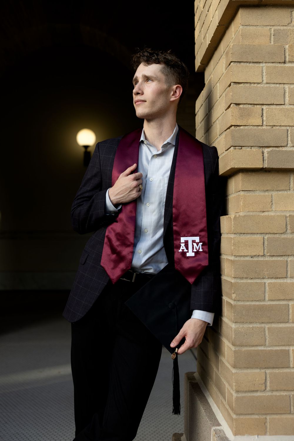 Young man in black suit wearing Texas A&M maroon graduation stole, leaning against brick wall in elegant senior portrait.