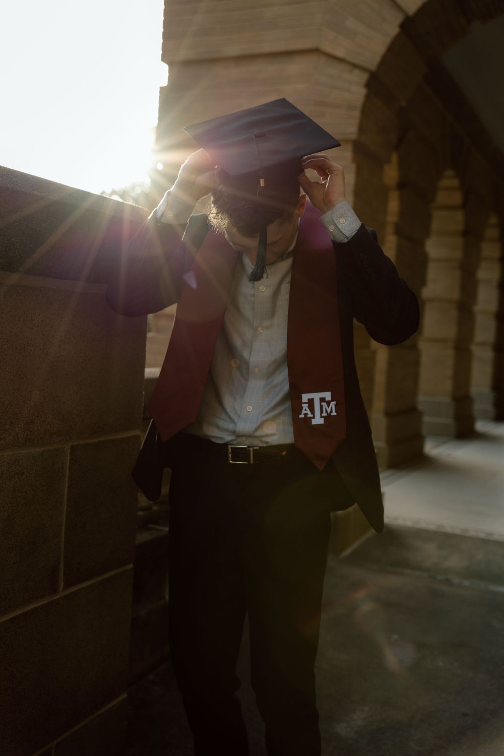 Texas A&M graduate adjusting cap in sunlight near stone archway, wearing maroon stole with ATM logo.
