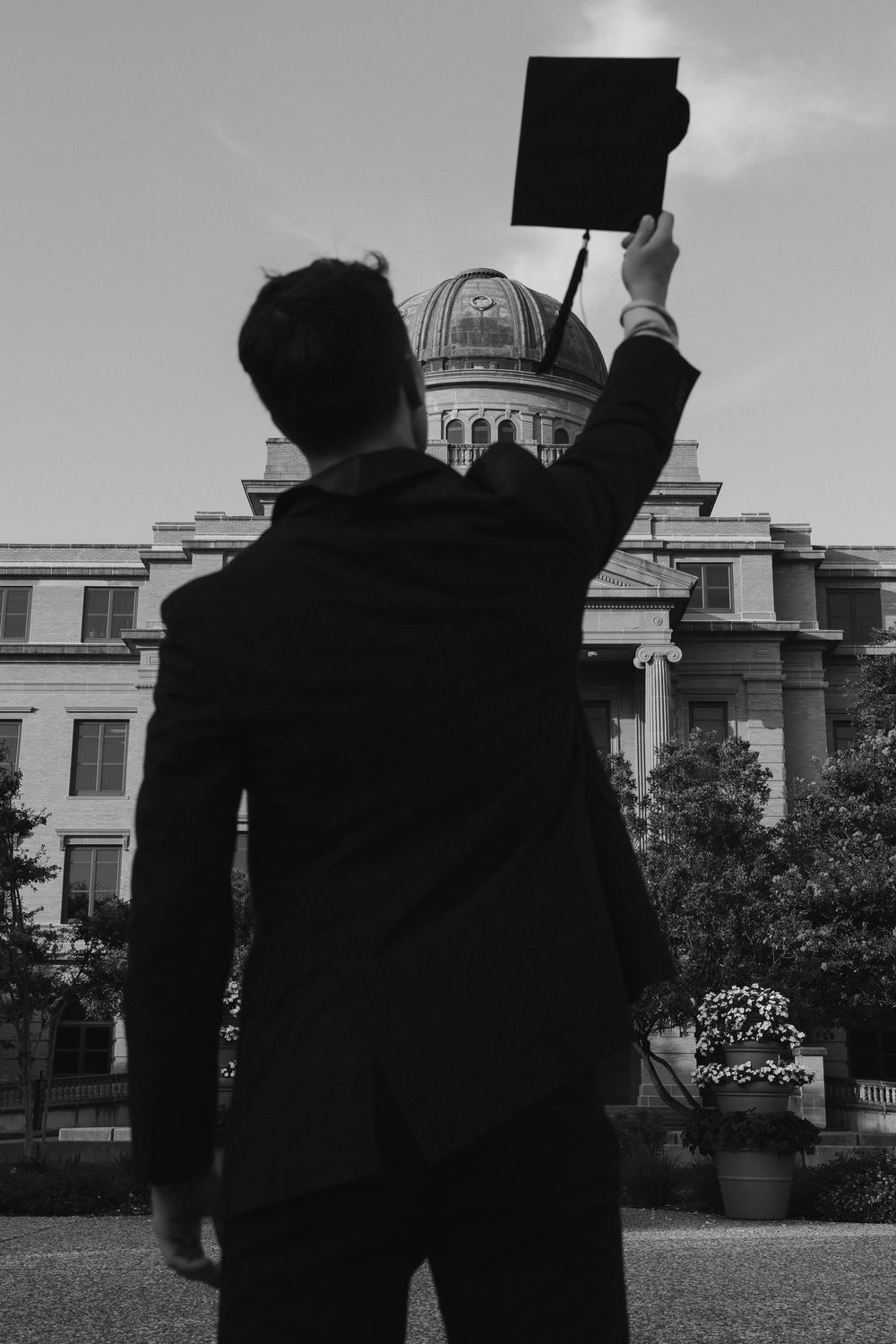 Black and white photo of a graduate raising his cap in front of a domed university building.