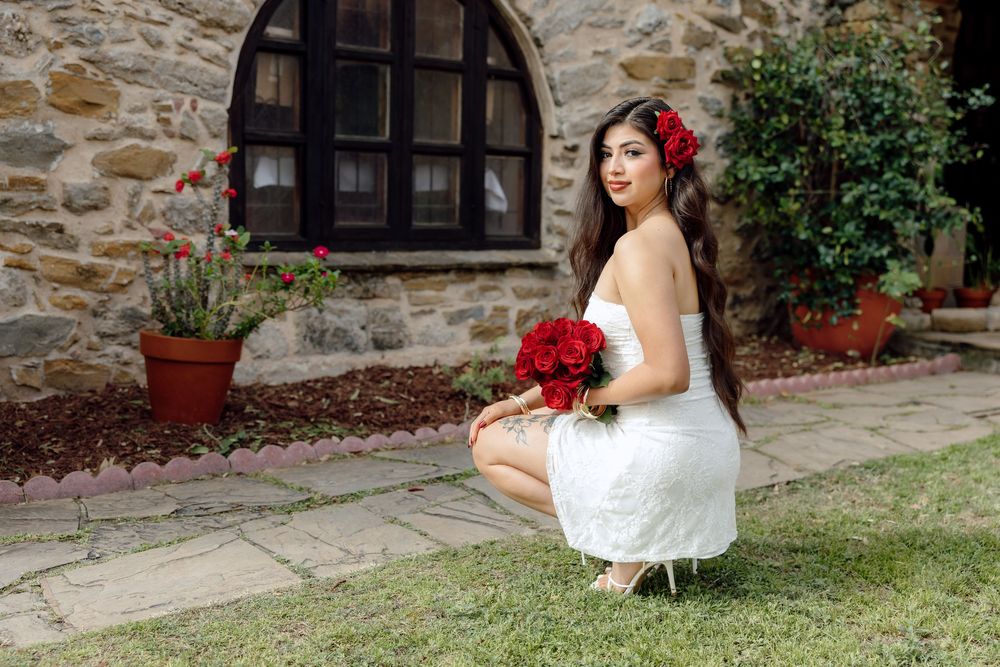 A bride in a white wedding dress poses with red roses against a rustic stone wall backdrop.