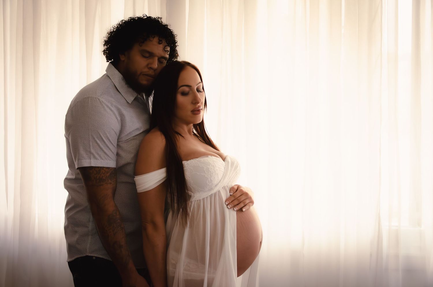 Intimate maternity photo of a couple embracing in soft window light.