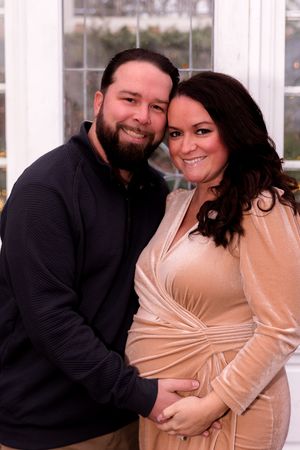 A couple shares an intimate moment in a maternity photoshoot featuring a beige wrap dress and black sweater.