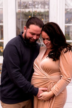Loving couple poses together during pregnancy photo session with white windows in background.