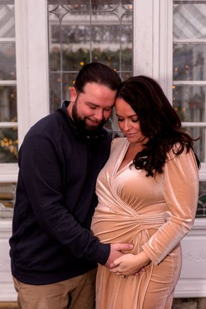 Tender maternity photography session showing couple embracing in front of bright windows.