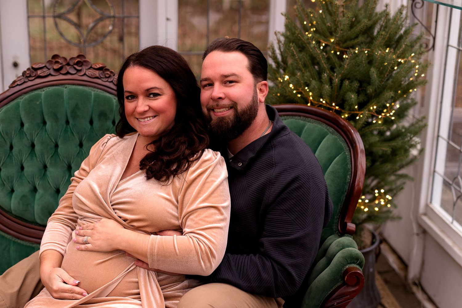 Expectant couple poses on green velvet couch with Christmas tree in background during photo session.