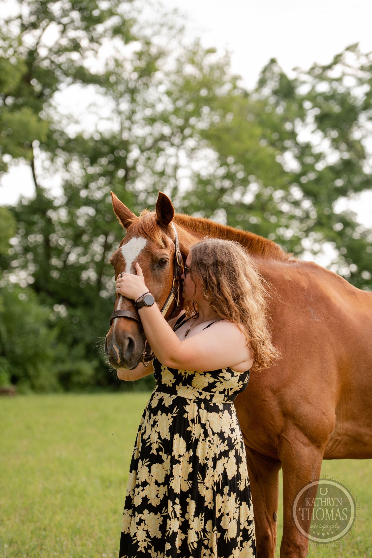 A chestnut horse with a white blaze stands close to someone in a floral dress outdoors.