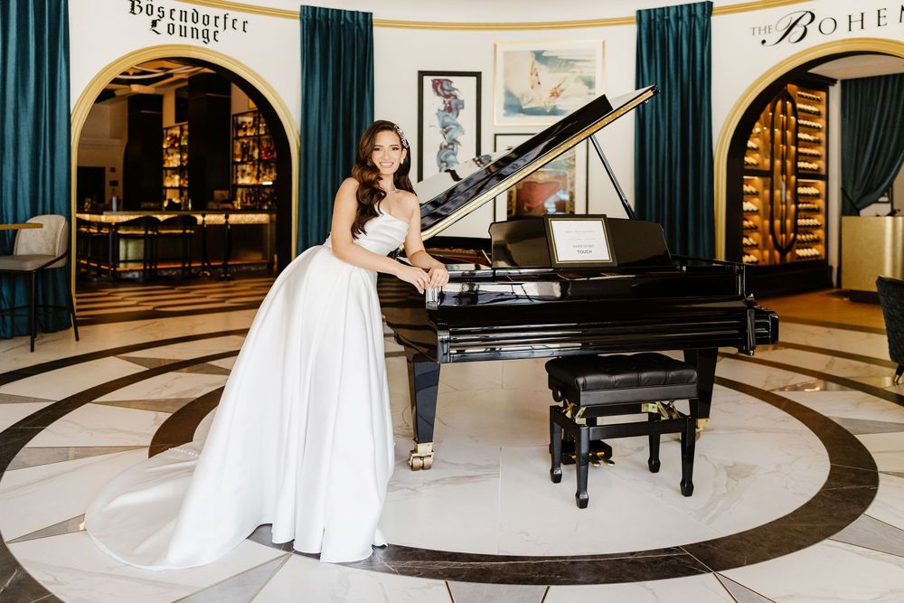 Bride in white gown posing elegantly beside a grand piano in an upscale hotel lobby with marble floors.