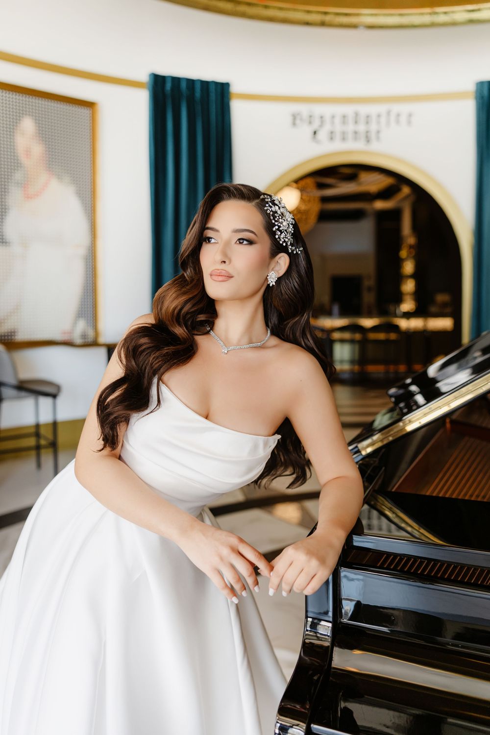 Elegant bride in strapless white gown with jeweled hair accessory poses beside a grand piano in a luxurious lounge.