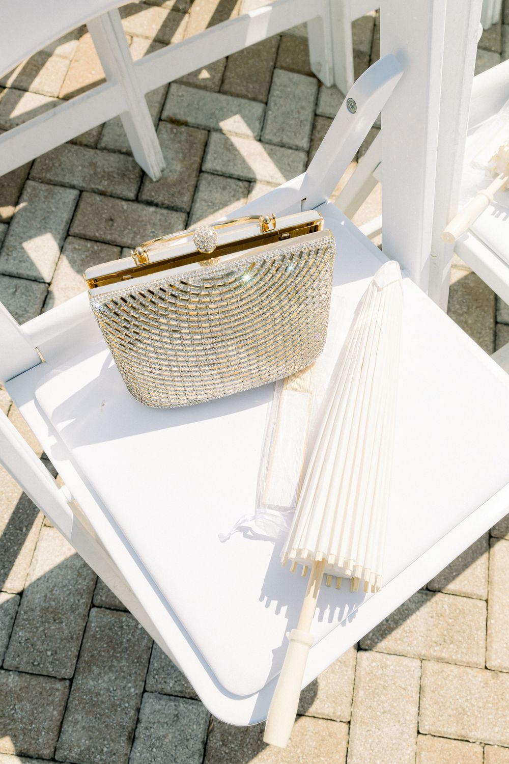 A vintage white wooden chair with a metallic clutch purse and parasol on a brick patio.