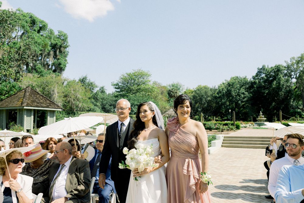 Wedding processional walking through sunny garden with decorative parasols and greenery.
