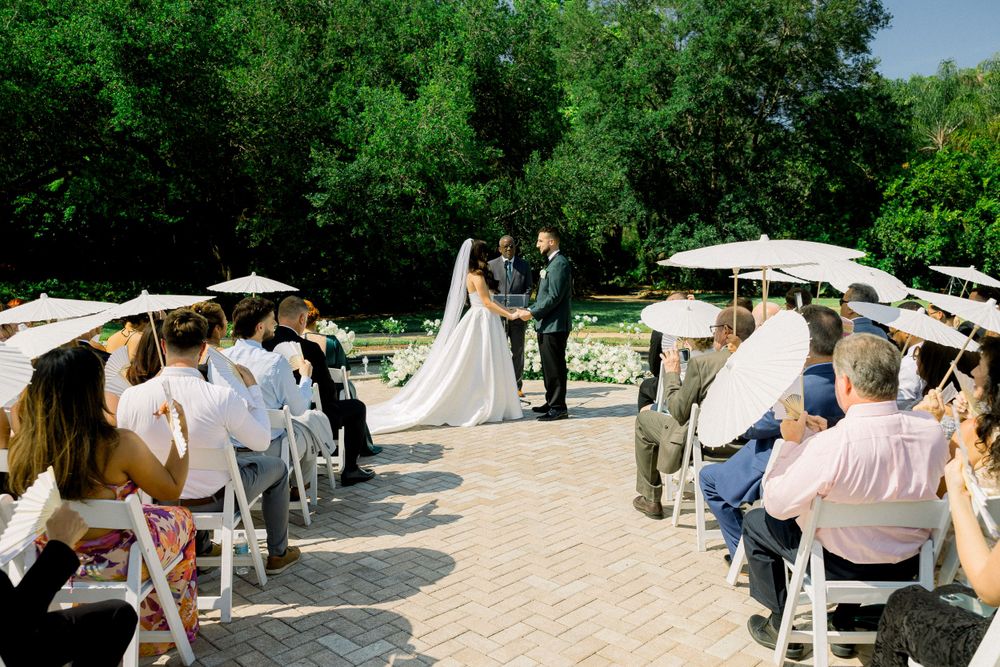 Wedding ceremony in progress with guests seated under white parasols in garden setting.