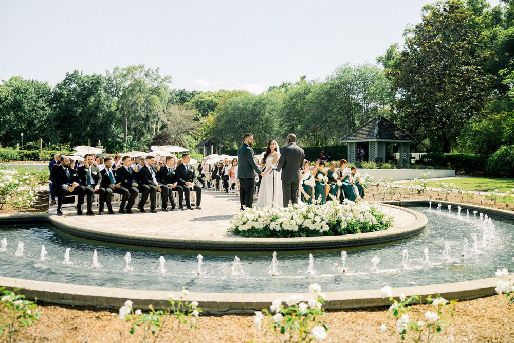 Wedding party gathered around circular fountain feature during outdoor garden ceremony.
