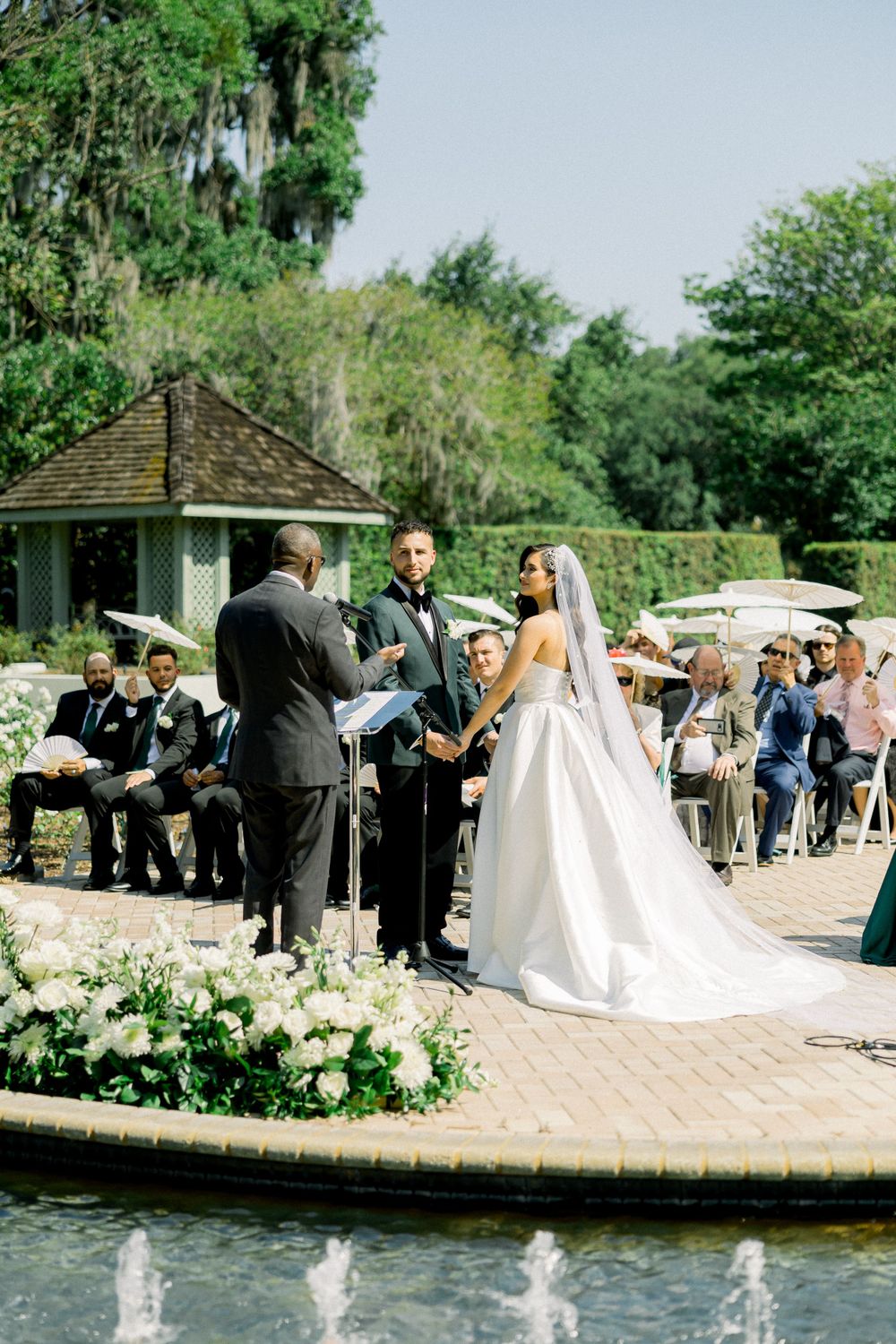 Wedding ceremony takes place on circular brick patio near fountain with garden views.