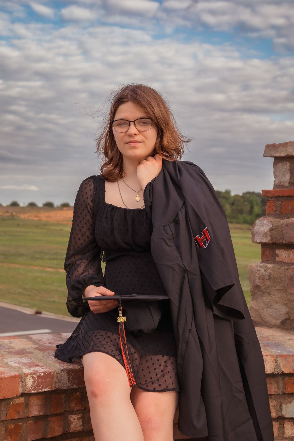 Graduate in black dress and academic gown sits on brick wall holding tablet during outdoor graduation photoshoot.